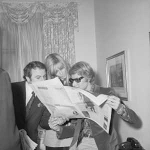 Yves Saint Laurent reading Women's Wear Daily with Pierre Berge and Betty Catroux after arriving in New York prior to attending a party given by Andy Warhol at the downtown Factory location, 1968.