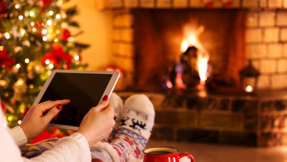 Woman sitting by fireplace and christmas tree holding tablet cozy concept