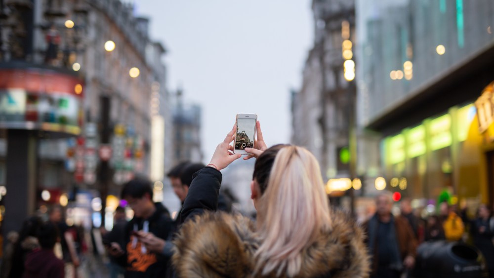 Woman tourist taking pictures of city with her cell phone on street in blurred background