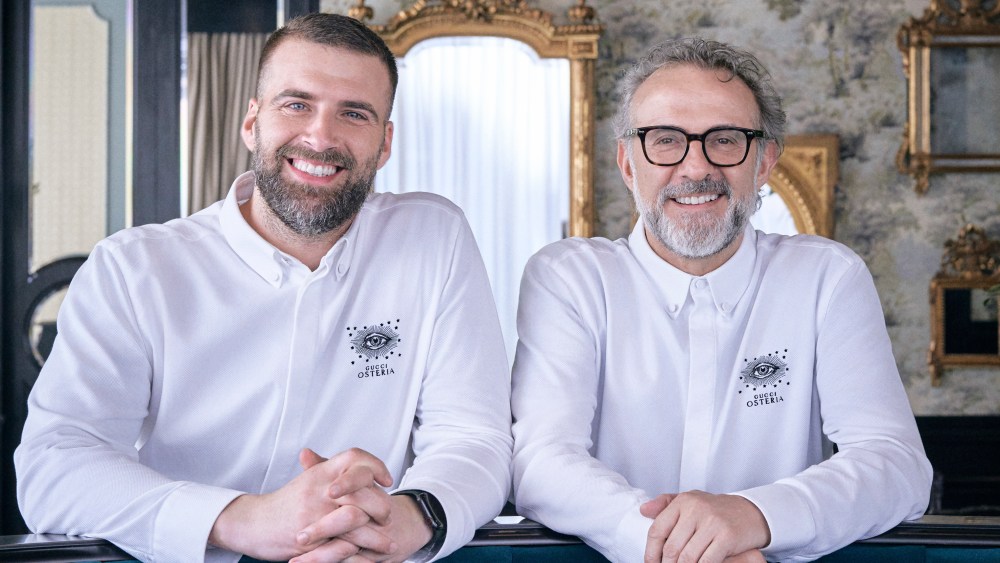 Antonio Iacoviello and Massimo Bottura pose smiling standing behind a blue quilted velvet couch, their arms resting on it.
