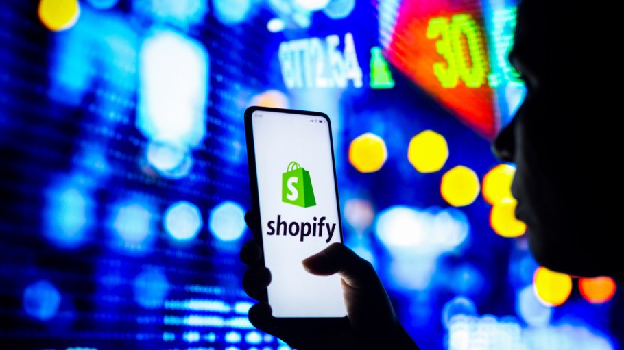 In this photo illustration, a silhouetted woman holds a smartphone with the Shopify logo displayed on the screen.