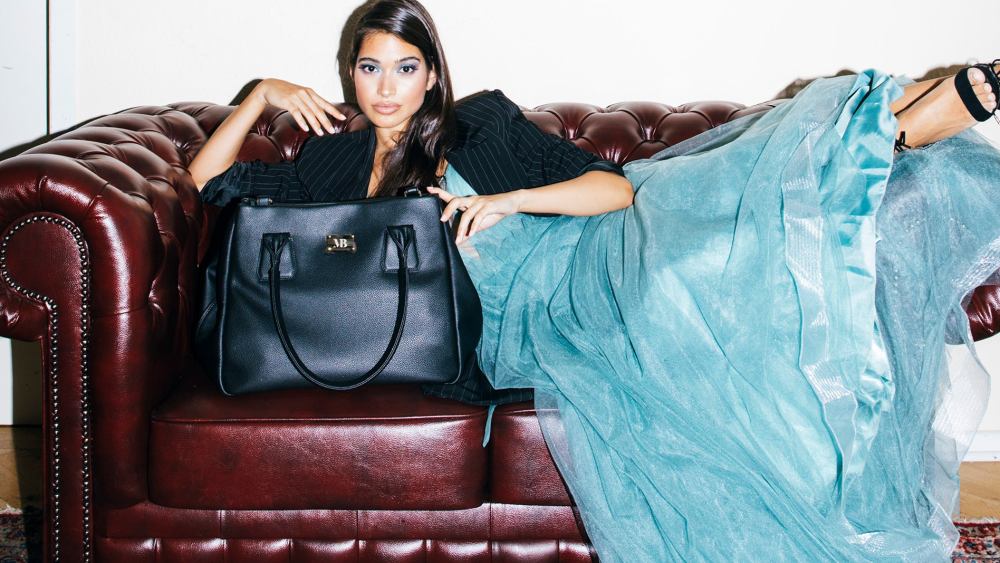 A black handbag style from Melina Bucher, a vegan luxury handbag brand, is displayed on a quilted couch with model posed in blue silky dress adjacent.