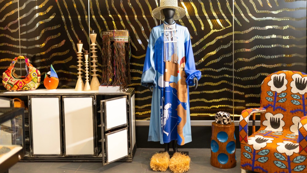 Alára shop at the Brooklyn Museum alongside the 'Africa Fashion' exhibit. Manikin wears blue dress with African women printed on the front and a straw hat, standing next to a decorative chair and wooden end table.