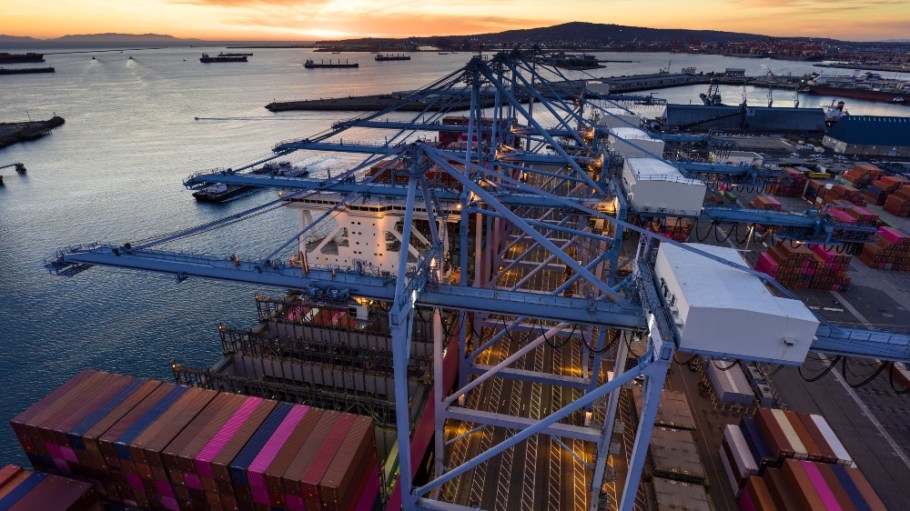 Aerial shot of a massive cargo ship loaded with containers docked in the Port of Long Beach, with the booms of ship-to-shore gantry cranes reaching out from the pier of the terminal.