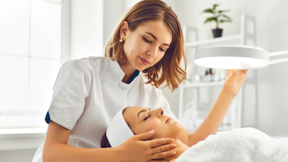 A cosmetologist or dermatologist directing lamp for facial treatment to young woman.